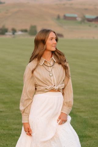 A woman stands on a green grassy field with rolling hills and distant buildings in the background, wearing a beige long-sleeve button-front top tied at the waist, a white midi skirt with eyelet lace, and brown knee-high boots.