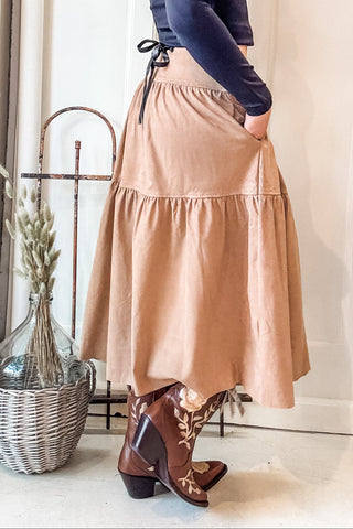 A woman stands in a doorway wearing a dark navy long-sleeve crop top and a beige tiered midi skirt with side pockets, paired with brown cowboy boots. The setting features a brick wall on the left, a white paneled door behind her, and a potted cactus on the right.
